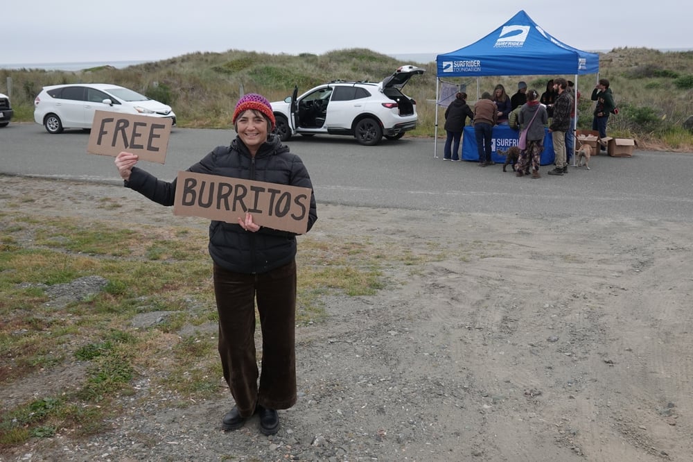 A woman holds a sign reading 'Free Burritos' in front of a blue Surfrider tent.