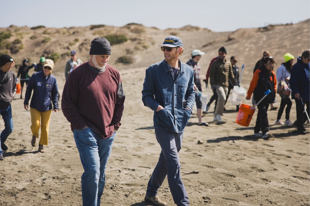 Two men walk on the beach with a crowd walking alongside.